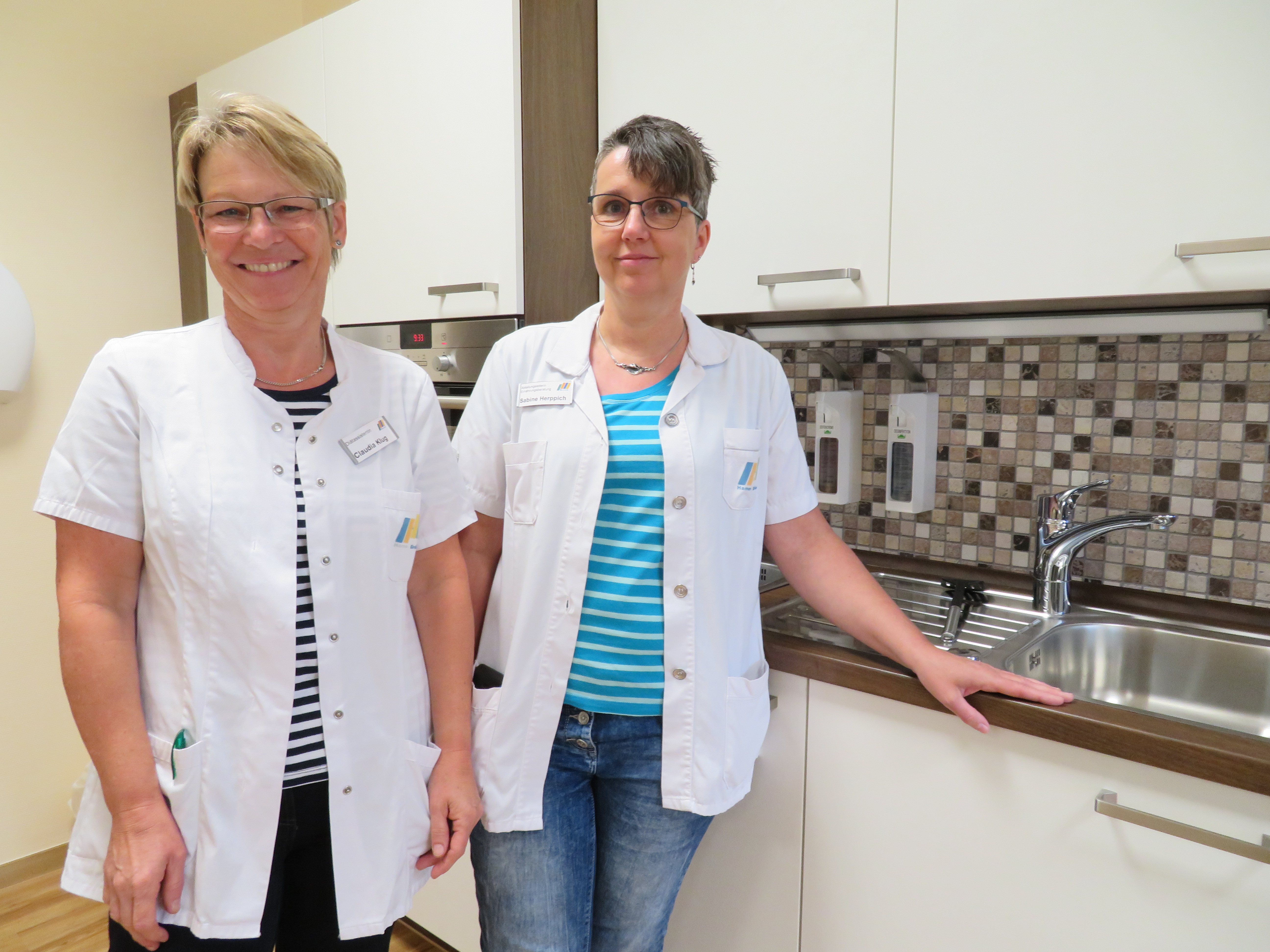 Zwei Frauen in weißen Arbeitsjacken stehen neben einer Küchenzeile mit Spüle und einem Fliesenrückwand.